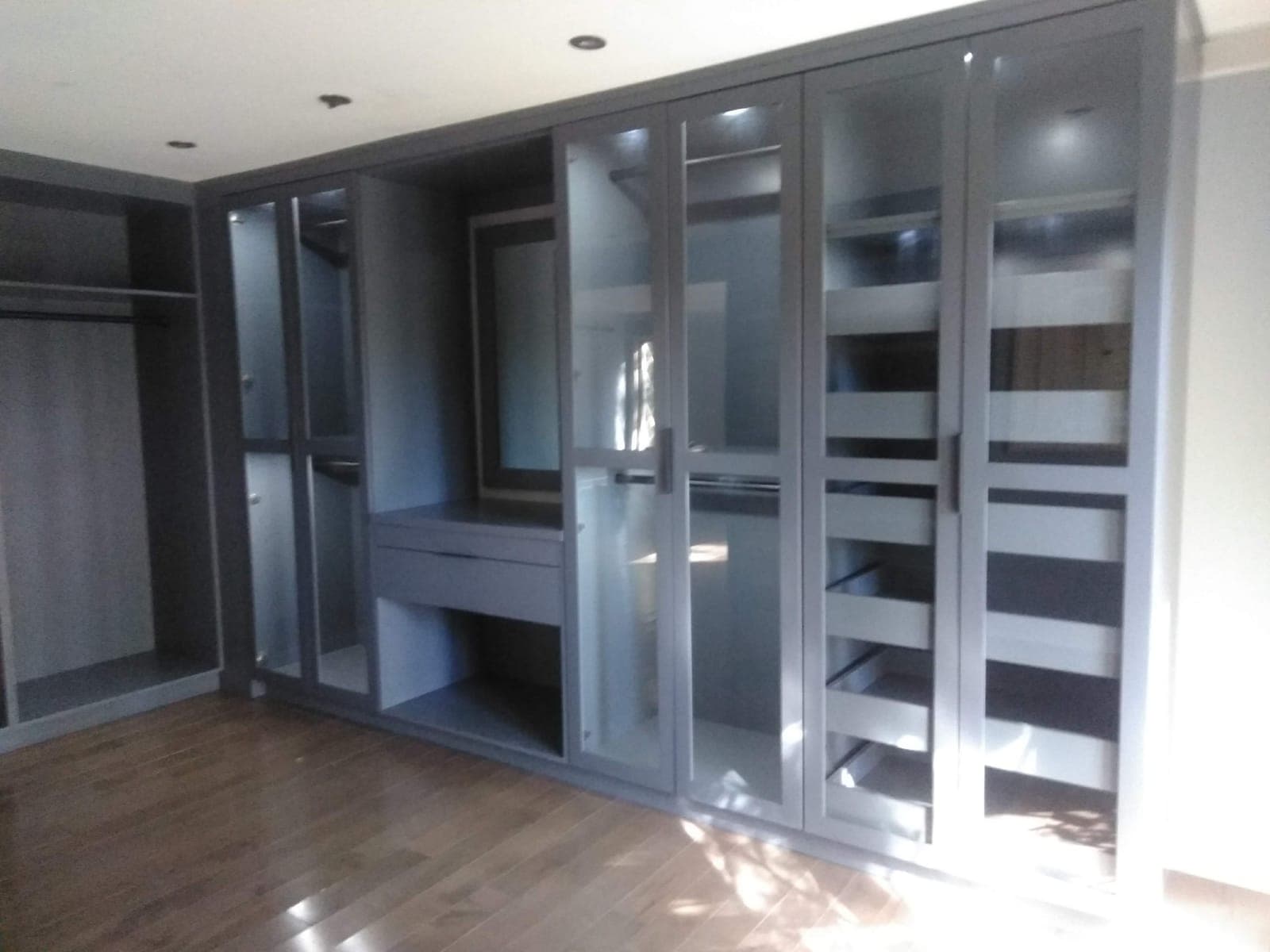 Gray built-in wardrobe wall with glass-front doors and interior lighting by Hdez Gabinets